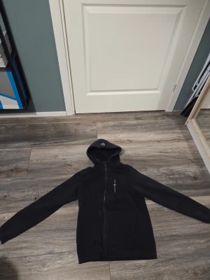 Svart hoodie från Sail Racing - Svart hoodie med dragkedja från Sail Racing. Hoodien har en bröstficka med dragkedja och logga tryckt på vänster sida. Tillverkad i mjukt material och har en klassisk huva. Perfekt för en avslappnad och sportig stil.