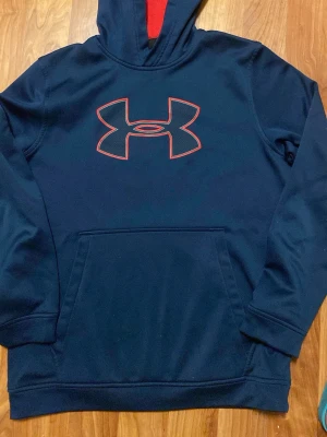 Mörkblå hoodie från Under Armour - Snygg mörkblå hoodie från Under Armour med stor röd logga på bröstet och röd detalj i huvan. Tröjan har en klassisk känguruficka och ribbade muddar vid ärmslut och nederkant. Perfekt för dig som gillar sportig stil eller vanlig stil och vill ha något bekvämt. Den har inga skador och är nästan helt ny.