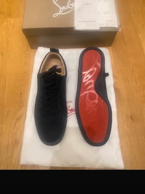 Mörk blåa sneakers från Christian Louboutin - Snygga mörk blå sneakers från Christian Louboutin med klassisk röd sula och diskret logga på hälen. Skorna har en låg profil, mörkblå skosnören och är tillverkade i mocka. Perfekta för dig som vill ha en exklusiv och stilren look.