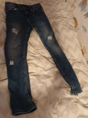 Blå slitna skinny jeans - Säljer ett par blå skinny jeans med slitningar och trasiga detaljer på benen. Jeansen har klassisk femficksdesign och är gjorda i denim. Perfekt för dig som gillar en trendig och avslappnad look.