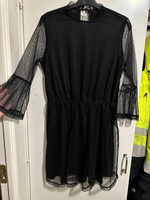 Svart meshklänning med volangärm strö: M - Snygg svart kortklänning i mesh med transparenta, prickiga ärmar och volangdetaljer vid ärmslutet. Klänningen har rund halsringning och markerad midja för en feminin siluett. Perfekt för dig som gillar trendiga detaljer och en luftig känsla. En knapp fattas baktil.