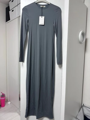 Grå långklänning från Pull&Bear - En stilren grå långklänning från Pull&Bear med rund halsringning och långa ärmar. Klänningen har en rak passform och är tillverkad i ett mjukt och stretchigt material som ger en bekväm känsla. Perfekt för dig som gillar minimalistisk stil.