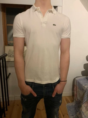 Vit pikétröja från Lacoste - Klassisk vit pikétröja från Lacoste med korta ärmar och den ikoniska gröna krokodilloggan på bröstet. Tröjan har krage och knappar vid halsen. Perfekt till jeans eller shorts för en clean och stilren look.