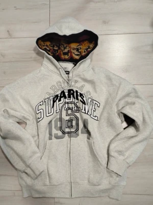 Supreme x Margiela grå hoodie - Grå zip-hoodie från Supreme och Maison Margiela med tryck 'SUPREME 1994 PARIS' på bröstet och coolt dödskallemönster i huvan. Mjuk bomullsblandning, två fickor fram och ribbade muddar. Perfekt för dig som gillar streetwear och unika detaljer.