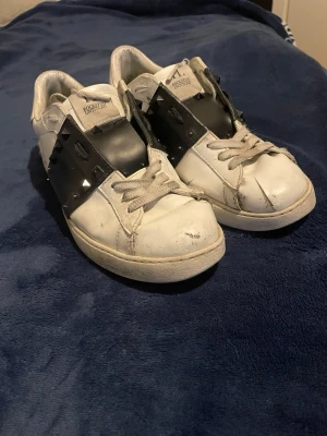 Valentino Garavani Rockstud sneakers - Snygga vita sneakers från Valentino Garavani med svart band över vristen dekorerat med nitar. Skorna har klassisk låg profil, vita snören och detaljerade sömmar. Tillverkade i läder för en lyxig känsla och edgy look. (Inte äkta Valentinos) (Väl använda)