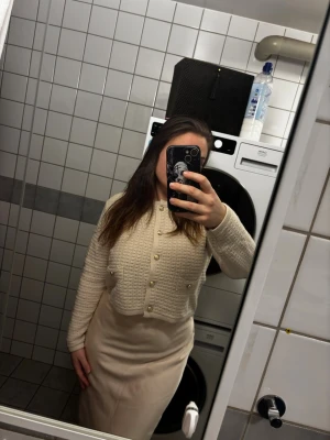 Krämvit kofta med pärlknappar - Säljer en krämvit, kort kofta med strukturerad stickning och dekorativa pärlknappar framtill. Koftan har långa ärmar och en klassisk rund halsringning. Perfekt för att ge din outfit en elegant touch.