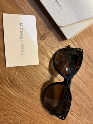 Svarta/bruna solglasögon från Michael Kors - Snygga svarta solglasögon från Michael Kors med rundad, kattögonformad båge och mörka linser. Bågen har små guldfärgade detaljer vid hörnen och är tillverkad i syntetmaterial. Perfekt för att ge din look en lyxig touch.