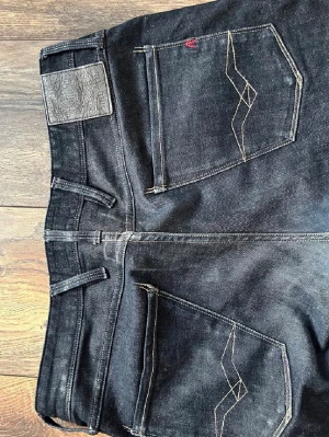 Svarta jeans från Replay, modell Anbass - Snygga svarta jeans från Replay, modell Anbass. Jeansen har raka ben, klassiska bakfickor med dekorativa sömmar och Replay-logga. Tillverkade i denim med en skön stretch för extra komfort. Perfekta för en trendig och avslappnad stil.