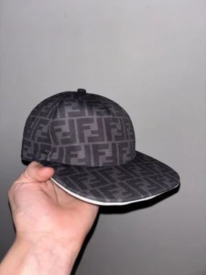 Svart keps med Fendi-logga - Snygg svart keps från Fendi med det klassiska FF-mönstret över hela kepsen. Kepsen har böjd skärm och är tillverkad i ett tygmaterial. Perfekt för dig som vill sticka ut med en lyxig accessoar.