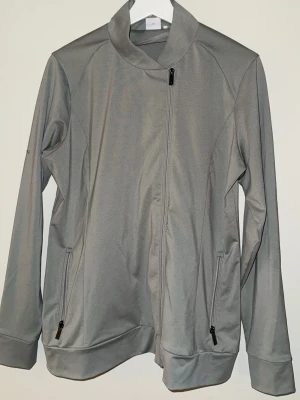 Grå sportjacka från Ping - Snygg grå sportjacka från Ping med asymmetrisk dragkedja och två fickor med dragkedja. Tillverkad i ett mjukt och stretchigt material, perfekt för träning eller golf. Jackan har hög krage och lång ärm för extra komfort.