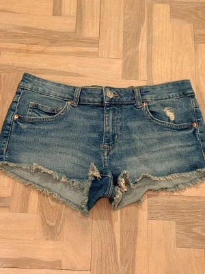 Blå jeansshorts med slitningar - Säljer ett par blå jeansshorts med råa, fransiga kanter och slitna detaljer framtill. Klassisk femficksmodell med knapp och dragkedja. Perfekta för sommaren och ger en avslappnad vibe.