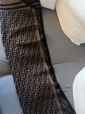 Brun och svart halsduk från Fendi - Snygg halsduk från Fendi i brunt och svart med det klassiska FF-monogrammet över hela tyget. Halsduken har en rektangulär form och är tillverkad i ett mjukt, stickat material som känns lyxigt och trendigt. Perfekt accessoar för att lyfta din outfit.