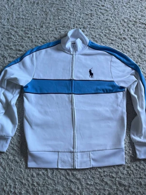 Vit och blå zip hoodie från Polo Ralph Lauren - Snygg vit hoodie med blåa detaljer och dragkedja från Polo Ralph Lauren. Klassisk logga på bröstet och blå rand längs ärmarna och bröstet. Tillverkad i mjukt material, perfekt för en avslappnad och sportig stil. Hög krage och ribbade muddar.