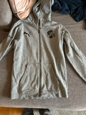 Grå zip hoodie från Puma - Snygg grå hoodie från Puma med dragkedja, huva och fickor framtill. Har svart Puma-logga på bröstet och klubbmärke på andra sidan. Tillverkad i mjukt bomullsmaterial, perfekt för chill eller träning. Diskret tryck på ärmen.