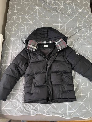 Svart pufferjacka med huva - Säljer en svart pufferjacka från Zara med stor huva och rutigt foder i grått, rött och vitt. Jackan har två dragkedjefickor framtill, tryckknappar och dragkedja. Perfekt för kalla vinterdagar och riktigt skön att bära.