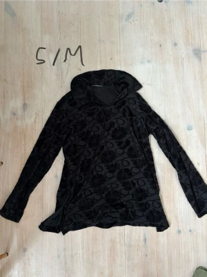 Svart mönstrad polotröja - Svart långärmad polotröja med blommigt sammetsmönster. Tröjan har en lös passform och hög krage som ger en avslappnad men ändå stilren look. 