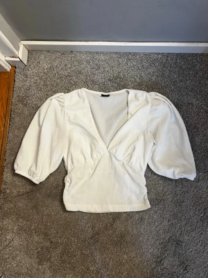 Vit v-ringad topp från Gina Tricot - Säljer en vit topp från Gina Tricot med v-ringning och puffiga trekvartsärmar. Toppen har en ribbad struktur och smickrande passform med lätt markerad midja. Perfekt att styla med både jeans och kjol.