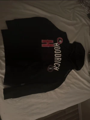Svart Hoodrich hoodie med tryck - Cool svart hoodie från Hoodrich med stora vita och röda tryck på bröstet. Hoodien har en klassisk huva med dragsko och flera broderade detaljer, bland annat ett stort H och rund logga. Perfekt för dig som gillar streetwear och vill sticka ut.