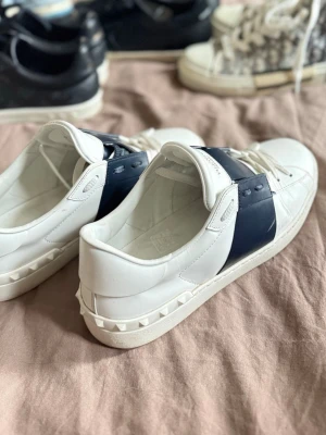 Valentino vita sneakers med blå detalj - Säljer ett par snygga vita sneakers från Valentino med bred mörkblå rem över mitten och coola nitar längs sulan. Skorna har klassisk låg profil, vita snören och logga på plösen. Perfekta för dig som vill ha en stilren men ändå unik look. Finns låda till. 