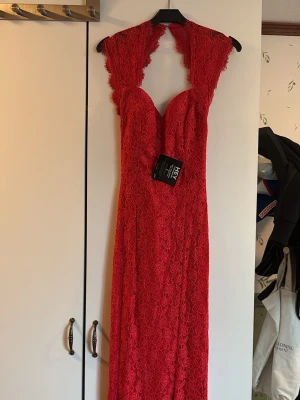 Röd lång spetsklänning från Nelly med v-ringning - Snygg röd långklänning med v-ringning och spetsdetaljer. Klänningen har ärmlös design och figurnära passform, perfekt för bal eller fest. Tillverkad i polyester med vackert blommigt spetsmönster och dragkedja i ryggen.