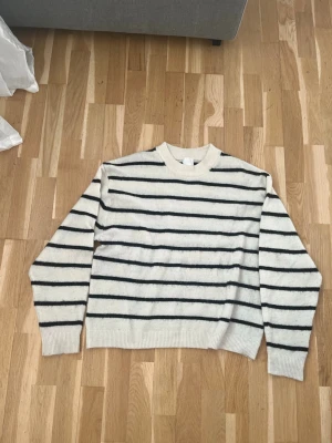 Vit stickad tröja med svarta ränder - Klassisk vit stickad tröja med svarta horisontella ränder och rund hals. Tröjan har långa ärmar och ribbstickade muddar vid ärmslut och nederkant. Perfekt för en avslappnad och stilren look.