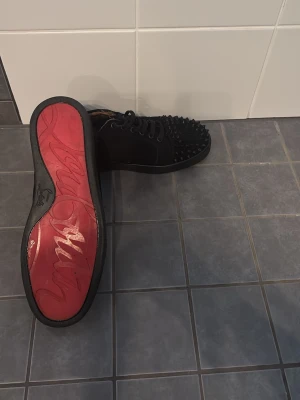 Louboutin skor - Säljer ett par svarta sneakers från Christian Louboutin med klassisk röd sula och svarta nitar över tån. Skorna har snörning och är tillverkade i mocka och läder för en lyxig känsla. Perfekta för dig som vill sticka ut med en edgy och exklusiv stil.