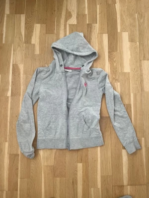 Grå hoodie med dragkedja från Cubus - Snygg grå hoodie från Cubus i storlek XS. Den har dragkedja framtill, två fickor och en huva. Rosa detaljer på insidan av kragen och ett litet rosa tryck på bröstet. Perfekt för chill dagar och enkel att matcha med jeans eller leggings.