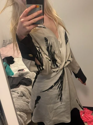 Blus i beige och svart - Snygg blus i beige med svart abstrakt mönster. Klänningen har långa ärmar med svarta muddar och guldknappar. Djup v-ringning och knyt i midjan ger en cool och trendig look. Perfekt för dig som gillar statement-plagg.