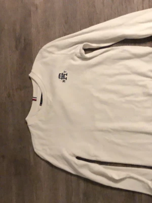 Vit sweatshirt från Tommy Hilfiger - Klassisk vit sweatshirt från Tommy Hilfiger med rund halsringning och diskret logga broderad på bröstet. Tröjan har långa ärmar och ribbade muddar vid ärmslut och nederkant. Perfekt för en clean och stilren look.
