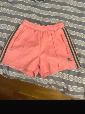 Rosa Adidas shorts med gröna ränder - Säljer ett par rosa Adidas shorts med gröna ränder längs sidorna och dragkedjefickor framtill. Shortsen har elastisk midja och Adidas-logga på vänster ben. Perfekta för sommaren och träning. nästan oanvända 