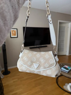 Beige axelremsväska från Guess - Snygg beige handväska från Guess med vit logotypmönster över hela väskan. Väskan har vita detaljer, kedjerem och ett trekantigt Guess-emblem framtill. Materialet är canvas med konstläderdetaljer och dragkedja baktill. Perfekt för dig som gillar trendiga accessoarer.