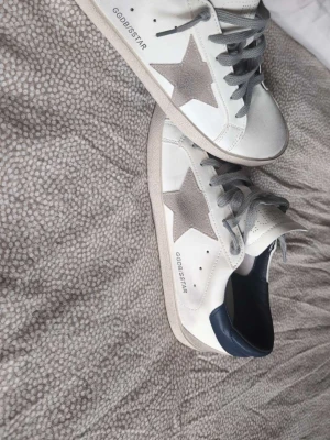 Golden Goose vita sneakers med stjärna - Golden Goose sneakers i vitt skinn med grå mockadetaljer och en ikonisk stjärna på sidan.  Skorna är storlek 42 men passar också 43✨ Skorna har grå snörning, blå hälkappa och platt sula. Kommer med original dustbag. Perfekta för dig som gillar en clean men ändå unik look. Reps