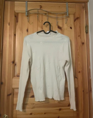 Vit ribbad stickad tröja med blommor - Säljer en vit ribbad stickad tröja med broderade blommor. Tröjan har långa ärmar, rund halsringning och vågig kant nedtill. Materialet är tunt och stretchigt, perfekt för lager på lager. Enkel men söt design som passar till det mesta.