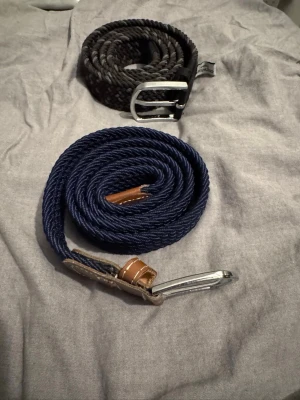 Flätade bälten - Säljer två snygga flätade bälten, ett i marinblått och ett i svart. Båda har silverfärgade metallspännen och detaljer i brunt skinn. Perfekta för att ge din outfit en cool touch. Bälten från one wear. Den blåa 100kr den andra 150kr, båda för 200