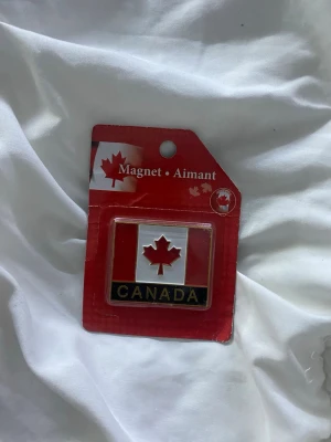Kanada-flagga kylskåpsmagnet - Säljer en rektangulär kylskåpsmagnet med Kanadas flagga och texten 'CANADA' i svart och guld. Magneten är röd, vit och svart och kommer i originalförpackning. Perfekt som souvenir eller dekoration till kylskåpet.