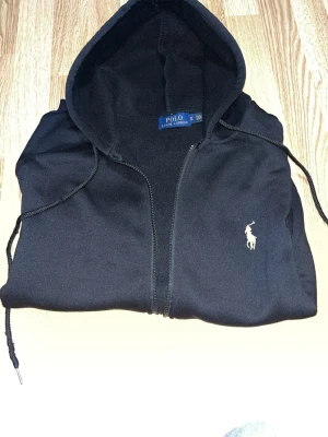 Svart hoodie från Polo Ralph Lauren -  PRIS KAN DISKUTERAS!!!!! Säljer en svart hoodie med dragkedja från Polo Ralph Lauren. Klassisk design med huva, dragsnören och fickor framtill. Ikoniska vita logon broderad på bröstet. Perfekt för en chill och stilren look. 
