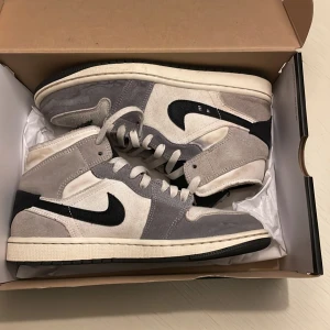 Nike Air Jordan 1 High grå/vit mocka - Nike Air Jordan 1 High sneakers i grå och vit mocka med svarta detaljer och klassisk svart swoosh. Skorna har högt skaft, perforerad tå och vita snören. Yttersulan är svart och ovandelen är tillverkad i en mix av mocka och textil för en clean och stilren look. Har använts drygt en vecka bara.