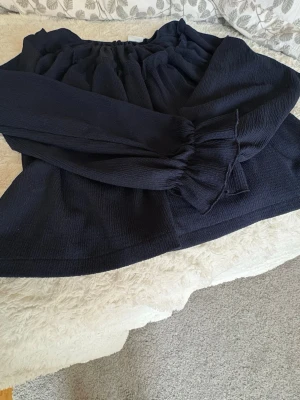 Mörkblå blus med volangärm - En snygg mörkblå blus med långärmade volangärmar detaljer upptill. Blusen har en lös passform och är tillverkad i ett lätt, strukturerat tyg som ger en trendig look. Perfekt att styla med jeans eller kjol för en cool vardagsstil. Kommer ej till användning.