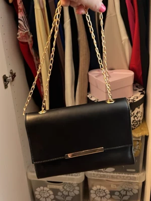 Svart kuvertväska med guldkedja - Stilren svart kuvertväska med guldfärgad kedja och metallspänne framtill. Väskan har en rektangulär form och är tillverkad i slätt konstläder. Perfekt för att bära det viktigaste och ger en elegant touch till din outfit.