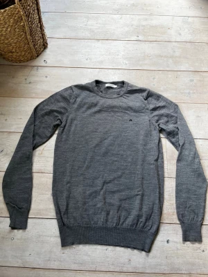 Grå merinoull stickad j lindeberg tröja  - Snygg grå stickad tröja med rund hals och diskret logga på bröstet. Tröjan har långa ärmar och ribbade muddar vid ärmslut och nederkant. Perfekt basplagg i mjukt material som passar till allt. Hör gärna av er vid minsta fundering😁