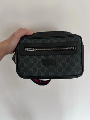 Svart axelväska från Gucci - Snygg svart axelväska från Gucci med klassiskt GG-mönster i grått. Väskan har en silverfärgad dragkedja framtill och justerbar axelrem i blått och rött. Tillverkad i slitstarkt canvasmaterial med detaljer i läder.