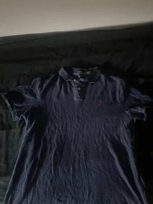 Marinblå pikétröja från Polo Ralph Lauren - Klassisk marinblå pikétröja från Polo Ralph Lauren med röd broderad logga på bröstet. Tröjan har krage, knappar framtill och korta ärmar. Perfekt för en stilren och avslappnad look.