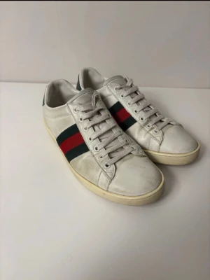 Gucci vita sneakers med röd/grön rand - Klassiska vita sneakers från Gucci med den ikoniska röd-gröna randen på sidan. Skorna har vita snören, rund tå och är tillverkade i läder. Perfekta för dig som vill ha en clean och tidlös look med lyxig känsla. Pris kan diskuteras. Storlek 45