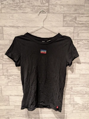 Svart Levi's t-shirt med logga - Svart t-shirt från Levi's med klassisk logga i rött, vitt och blått på bröstet. T-shirten har rund halsringning och korta ärmar. Tillverkad i mjuk bomull, perfekt för en avslappnad stil.