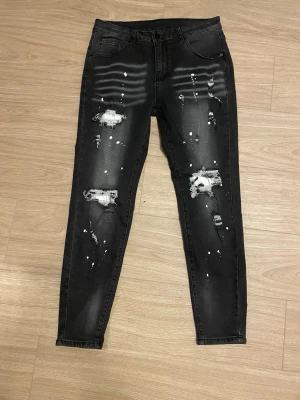 Helt ny svarta jeans med slitningar - Svarta skinny jeans med kraftiga slitningar och vita färgstänk på både fram- och baksida. Jeansen har klassisk femficksmodell och smal passform. Perfekta för en edgy streetstil.Storlek M. Helt ny.