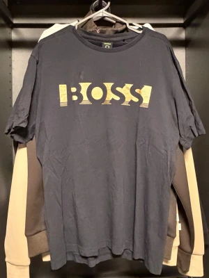 Svart BOSS t-shirt med gul logga - Snygg svart t-shirt från BOSS med gul logga i grafiskt tryck på bröstet. Klassisk rund hals och korta ärmar. Tillverkad i mjuk bomull för skön känsla och enkel stil. Perfekt för dig som gillar stilrena och trendiga plagg.