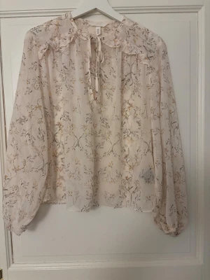 Blommig transparent blus med volang - Typ aldrig använd, Söt blus från H&M i ljusbeige/ljusrosa transparent tyg med blommigt mönster i dova färger. Blusen har volang vid axlarna, knytband vid halsen och långa, luftiga ärmar med resår vid handlederna. Perfekt för en romantisk och trendig look.