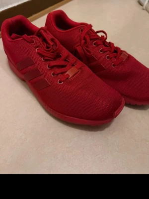 Röda Adidas sneakers i mesh - Snygga helröda sneakers från Adidas med klassiska tre ränder på sidan. Skorna är låga och har ovandel i ventilerande meshmaterial samt matchande röda snören och sula. Perfekta för en sportig och trendig look.