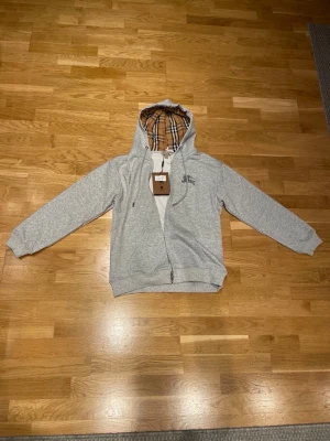 Grå hoodie från Burberry med dragkedja - Snygg grå hoodie från Burberry med dragkedja och klassiskt rutigt foder i huvan. Märkets logga är broderad på bröstet och luvan har justerbara snören. Tillverkad i mjukt bomullsmaterial för en bekväm känsla. Perfekt för en avslappnad och stilren look.