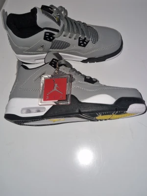 Gråa Air Jordan 4 Retro sneakers - Snygga Air Jordan 4 Retro sneakers i grått med svarta och vita detaljer samt gula inslag på sulan. Skorna har meshpaneler på sidorna, klassisk Jumpman-logga på plösen och en synlig Air-enhet i hälen. Tillverkade i läder och textil med robust yttersula.